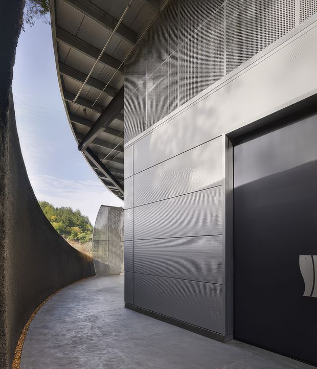 A close architectural exterior view of a contemporary building along a curved concrete walkway. The facade combines dark grey ribbed metal panels, a perforated metal screen at upper level, and a large matte-black door. A broad projecting roof with exposed steel framing creates a strong cantilevered edge, while a tall curved dark retaining wall encloses the path.