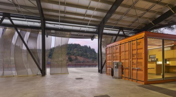 A large semi-open industrial interior with a polished concrete floor, exposed dark steel structure, corrugated metal roof, translucent hanging curtain panels, and an orange shipping-container-like enclosed unit with glazed openings and warm interior lighting. A wooded landscape is visible beyond the open sides.