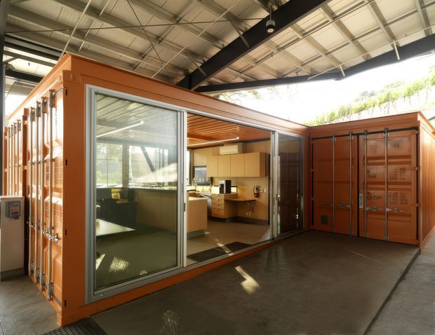 A converted shipping-container-style workspace or small commercial interior set within a large industrial warehouse. Orange-painted modular container walls frame expansive aluminium-framed sliding glass doors, revealing a compact kitchenette and office area with light timber cabinetry and dark work surfaces. The surrounding warehouse has exposed steel beams, a high corrugated metal roof, and polished concrete flooring. Bright daylight enters from the open warehouse side and through the glazing.