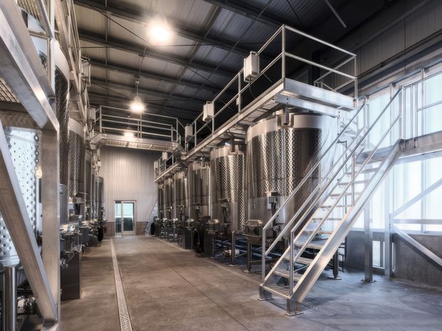 Industrial-scale beverage production or brewing facility interior with rows of tall stainless-steel cylindrical tanks, elevated metal walkways and platforms, access stairs, exposed structural framing, corrugated metal wall and ceiling surfaces, concrete flooring, and extensive translucent glazing on the right. The space is utilitarian, clean, and brightly lit.