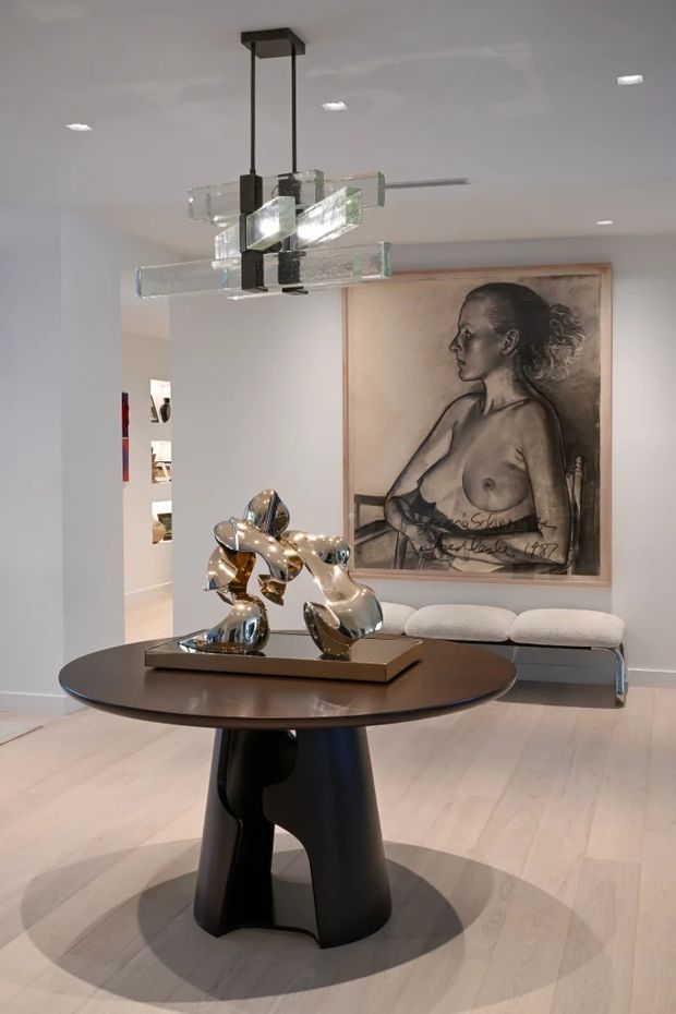A refined contemporary interior featuring a round dark timber or dark-stained table with a sculptural black pedestal base, a polished metallic abstract sculpture, a large framed portrait artwork, and a pale upholstered bench. A geometric suspended glass-and-metal chandelier provides a focal point above the table. The space has white walls, pale timber flooring, recessed ceiling lights, and an art-focused gallery-like atmosphere.