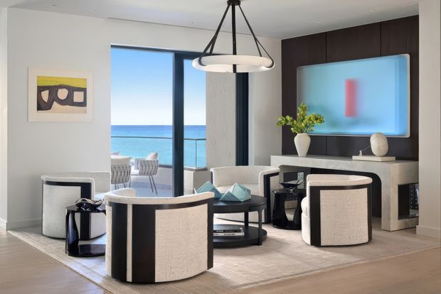 Contemporary living room overlooking the ocean, furnished with curved cream-and-black lounge chairs, a low coffee table, a pale sofa, and a sculptural suspended pendant light. A large wall-mounted screen is set into dark wood cabinetry above a stone or light-toned media console, with floor-to-ceiling glazing opening to a balcony and sea view.