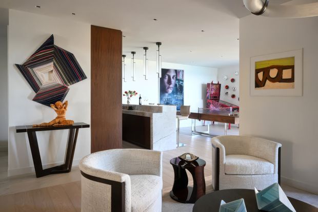 Refined open-plan contemporary interior combining a living area with a visible kitchen and dining space. The room features sculptural cream boucle-style armchairs, a dark console table, abstract wall art, a tall timber panel, stone kitchen surfaces, and minimalist pendant lighting. The palette is light and neutral with dark and colourful artistic accents.