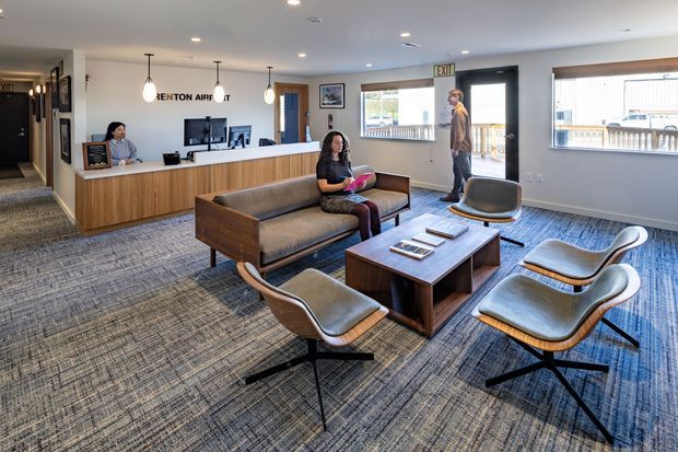 A bright, contemporary airport reception and waiting lounge with a long timber-fronted reception desk, monitor workstations, pendant lights, a mid-century-inspired sofa and lounge chairs, and a central timber coffee table. Large windows and glazed doors provide views outside, while patterned commercial carpet covers the floor.