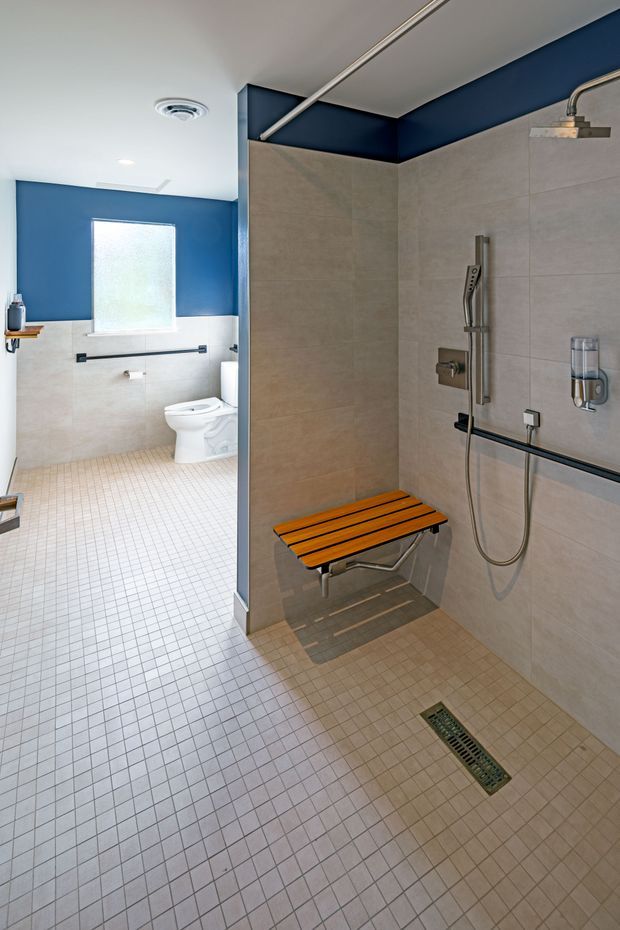 Accessible bathroom with a large open shower area, wall-mounted handheld shower, fold-down wooden shower seat, grab rails, tiled floor with linear drain, and adjacent toilet zone. The room combines light beige wall and floor tiles with a bold deep blue painted upper wall and white ceiling.