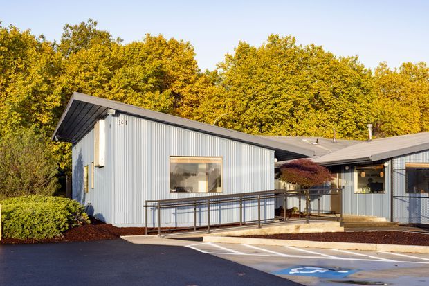Single-storey low-pitched commercial building with light vertical metal cladding, dark trim and roof, accessible ramp and handrails, and a covered entry. The building sits beside landscaped planting and a paved parking area with an accessible parking marking, with mature trees behind it.