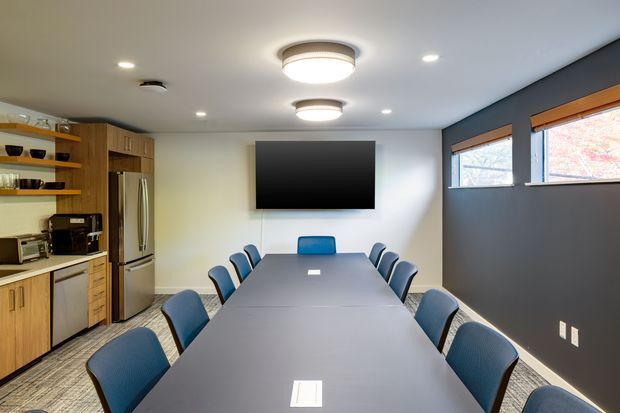 A modern meeting or dining room dominated by a long rectangular table with blue upholstered chairs. A wall-mounted flat-screen display faces the table, while a compact kitchenette with timber cabinetry, open shelving, stainless-steel refrigerator and appliances occupies the left side. The room has dark grey and white walls, carpet flooring, recessed and circular ceiling lighting, and narrow horizontal windows.