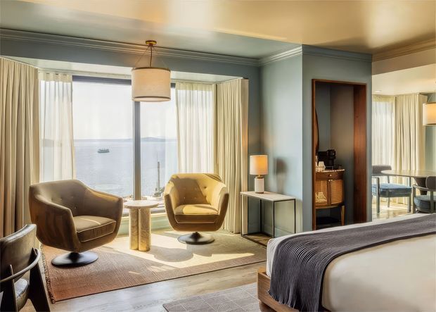 A refined bedroom with a mid-century-inspired sitting area overlooking water through expansive corner glazing. The room features two sculptural upholstered lounge chairs, a small round side table, a pendant light, a console cabinet, layered rugs, blue-grey painted walls with decorative ceiling trim, and a partially visible bed with a dark textured throw. A mirrored wall reflects additional seating and the waterfront view.