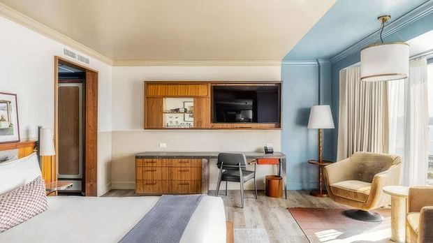 A well-appointed bedroom with a mid-century-inspired built-in timber entertainment and study wall, a large bed in the foreground, and a blue-painted sitting area by tall windows. The room features warm timber joinery, a compact desk, upholstered lounge chair, floor lamp, pendant light, patterned rug, and light timber flooring. The overall composition is refined and comfortable, with a blend of original architectural detailing and contemporary furnishings.