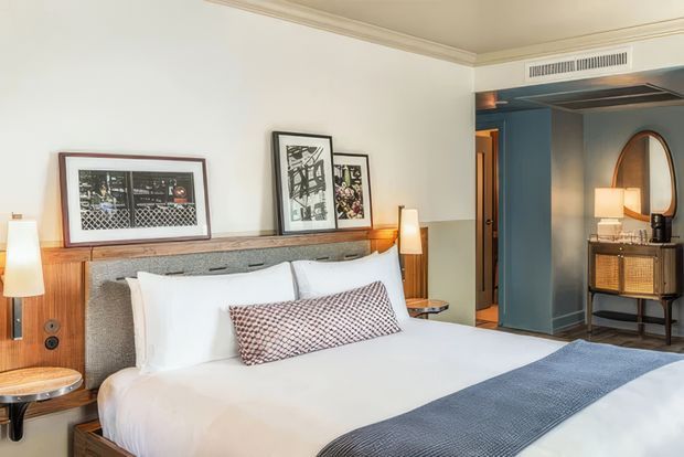 A warmly styled bedroom with a large bed, white bedding, a blue-grey runner, and a textured grey upholstered headboard integrated with timber bedside shelving and wall-mounted lamps. Framed artwork rests on the timber ledge above the bed. The room opens toward a dark blue hallway or adjoining area with a mirrored console and lamp visible through the doorway. Decorative ceiling trim and a linear air-conditioning vent add architectural detail.