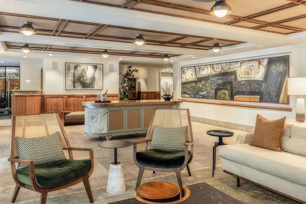 A refined hospitality-style lounge and reception area with mid-century-inspired timber and upholstered seating arranged around small side tables. A timber-fronted reception or bar counter sits centrally beneath a decorative exposed ceiling grid with warm pendant lighting. The space features light walls, polished stone flooring, a large framed artwork, timber joinery, and a softly neutral palette with green and rust-toned accents.