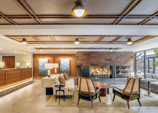 Spacious, warmly styled living room with a central fireplace clad in dark textured tile or brick, surrounded by timber cabinetry and wall panelling. The room features mid-century-inspired lounge chairs, a cream sofa, side tables, lamps, tiled flooring, expansive glazing, and a distinctive timber ceiling grid with exposed beam-like detailing. Multiple pendant and ceiling lights create a refined, inviting atmosphere.