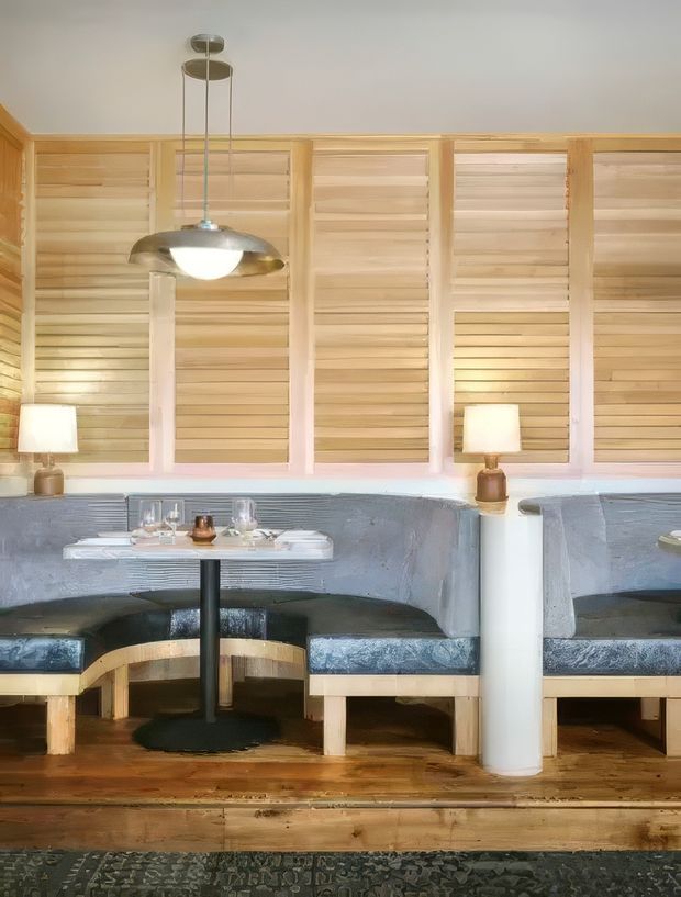 A warmly lit dining or lounge area with built-in curved banquette seating upholstered in dark blue-grey fabric, pale timber slatted wall panels, small round dining tables, table lamps, and a suspended metallic pendant light. The space has timber flooring and a softly textured, handcrafted interior with a relaxed contemporary feel.