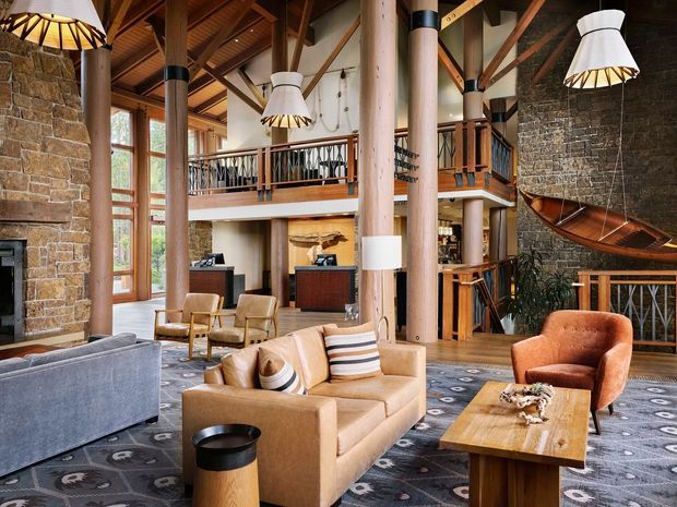 A spacious lodge-style living room with a dramatic double-height timber structure, exposed rafters, large natural-stone fireplaces or feature walls, timber columns, mezzanine-level balconies, and tall glazed doors. The room is furnished with leather and fabric sofas, armchairs, a timber coffee table, patterned carpet, and distinctive hanging pendant lights. A sculptural suspended boat-like feature hangs near the stone wall.