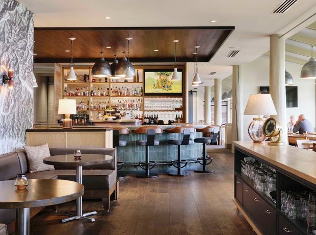 A sophisticated hospitality bar and lounge interior with a long central bar, green upholstered stools, extensive bottle and glass shelving, pendant lighting, and dark timber ceiling detailing. Lounge seating and small round tables occupy the foreground, while a dining area is visible beyond. The space combines warm timber, patterned wall covering, dark cabinetry, and polished decorative finishes in a refined contemporary setting.