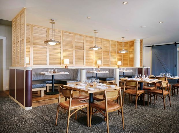 A refined restaurant dining room with multiple timber dining tables and curved-back wooden chairs arranged on a patterned carpet. Along the rear wall are upholstered booth seats beneath a full-height timber louvred feature partition, with marble-look tabletops, pendant lights, recessed ceiling lighting, and a large blue-grey sliding door. The space has a warm, contemporary hospitality interior with layered seating and custom joinery.