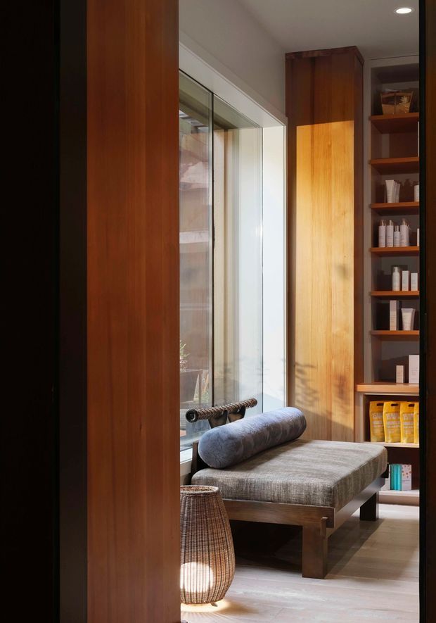 A warm, refined interior vignette featuring a timber-framed upholstered chaise or daybed beside full-height glazing. Floor-to-ceiling dark timber cabinetry and open shelving display bottles and toiletries, suggesting a dressing or bedroom-adjacent relaxation area. Natural wood, grey upholstery, woven lighting, and soft sunlight create a calm, tactile atmosphere.