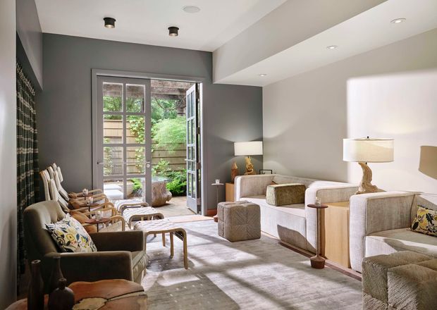 A bright, refined living room with two large cream upholstered sofas, dark accent armchairs, a pale area rug, and a pair of side tables with lamps. A full-height glazed door opens to a leafy outdoor area, while a dark grey feature wall and recessed ceiling lighting provide contrast. The room has a comfortable, layered seating arrangement and abundant natural light.