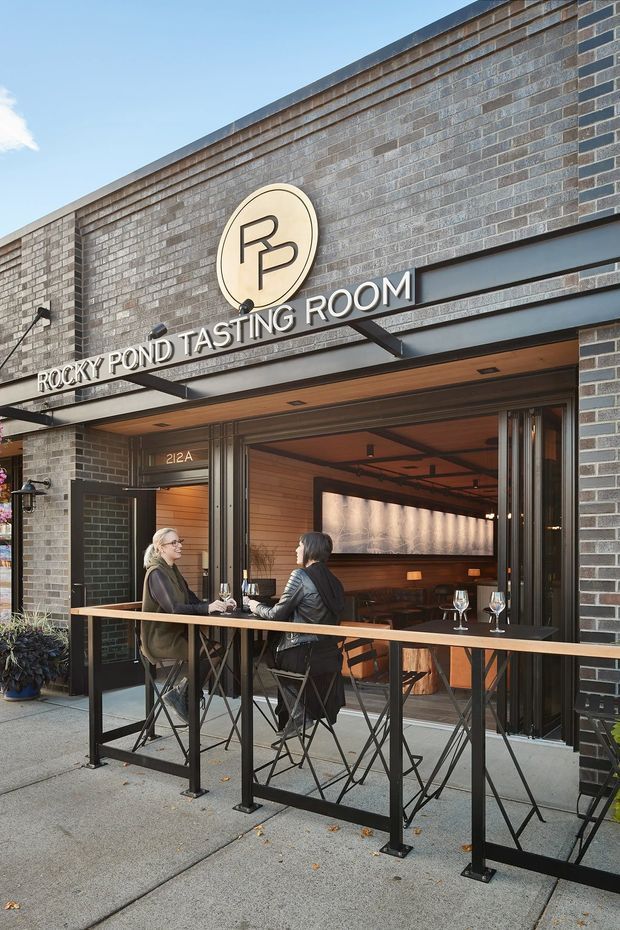 Exterior street-level view of the Rocky Pond Tasting Room, with a dark brick facade, prominent illuminated-style signage, open bi-fold or folding glass frontage, timber-lined soffit and interior ceiling, and an outdoor bar-height seating rail. Two people are seated at the exterior counter while the tasting-room interior is visible behind them.