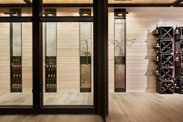 A refined wine storage and display area viewed through black-framed glazed doors. The room features light horizontal timber wall panelling, pale timber flooring, integrated vertical wine displays, and a dark geometric wine rack holding bottles on the right. Warm concealed lighting and dark metal framing create a sophisticated cellar-like atmosphere.