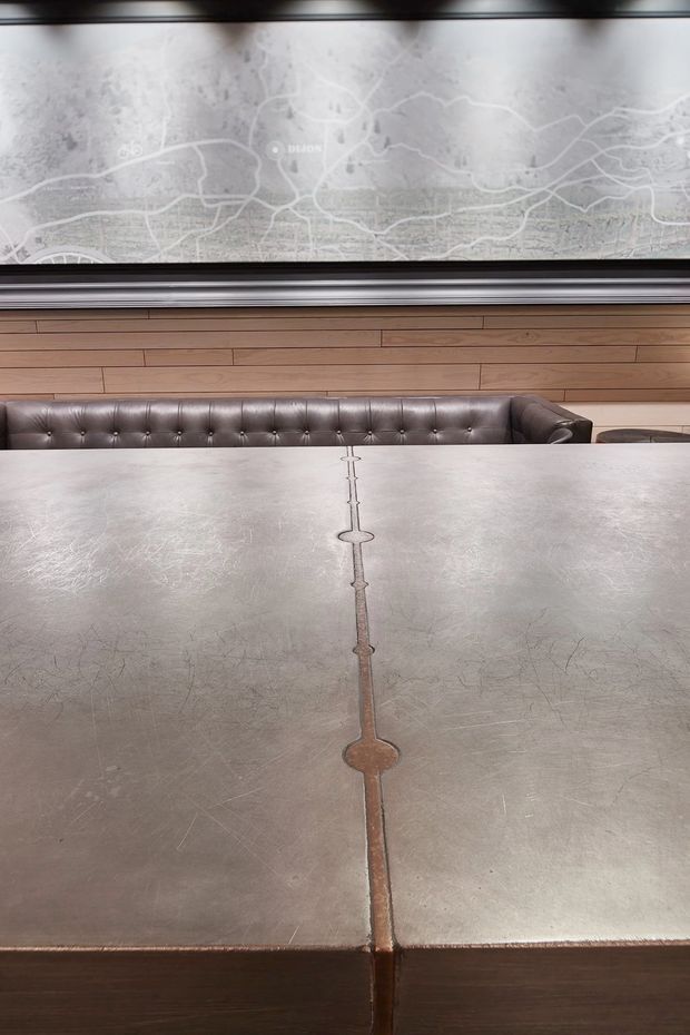 A close-up view of a long communal table with a weathered grey metal tabletop and a distinctive central brass-toned inlay or seam. Behind it is a dark brown tufted leather banquette, while the wall is clad in horizontal warm timber boards. A large monochromatic city map artwork or graphic spans the upper wall, framed by dark trim. The scene has a refined industrial hospitality or commercial-interior character.