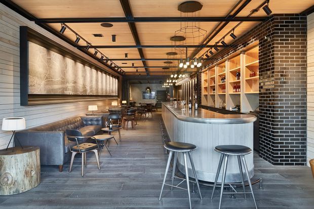 A long, sophisticated commercial bar and lounge interior with a curved brushed-metal bar, dark stools, open timber shelving, and a black brick feature wall. The space combines timber ceilings and flooring with grey horizontal wall cladding, leather banquette seating, lounge chairs, small tables, track lighting, pendant lights, and a large framed wall artwork. The design has an industrial yet refined hospitality atmosphere.