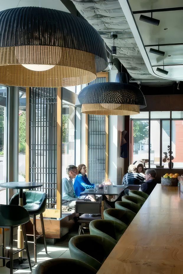 A contemporary café or hospitality interior with a long polished counter lined with dark green upholstered bar chairs. Large sculptural pendant lights made from dark ribbed and natural woven materials hang over seating areas. Floor-to-ceiling glazing, black metal screens, exposed ceiling services, and a mix of timber, concrete, and dark finishes create a refined urban atmosphere.