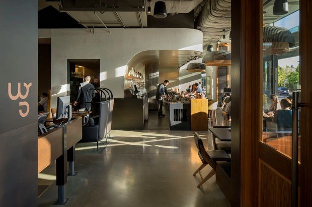 A sunlit contemporary café or restaurant interior viewed from the entrance, with a polished concrete floor, curved plaster feature wall and ceiling, exposed services overhead, timber-framed glazing and doors, bar service counter, banquette seating, tables, and patrons. The space combines industrial ceiling infrastructure with refined hospitality fit-out and strong afternoon light.