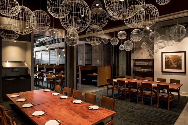 A large, warmly lit restaurant dining space with several solid timber communal and dining tables, dark upholstered seating, and an open kitchen at left. The industrial-inspired interior features dark structural columns, framed artwork, shelving, exposed ceiling services, and a striking installation of numerous suspended wire globe lights across the ceiling. A patterned dark carpet and timber furniture create a refined, intimate atmosphere.