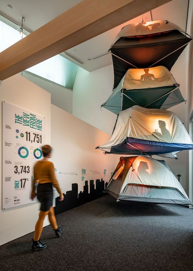 A tall, contemporary exhibition or commercial interior with a suspended vertical stack of illuminated camping tents forming the main installation. The space has white walls, dark carpet flooring, exposed light timber beams, a skylight or clerestory opening, and large graphic information panels with statistics along the wall. A blurred visitor walks through the display area.