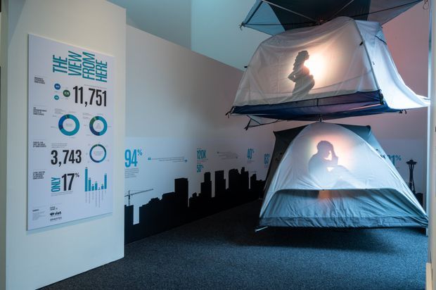 An indoor exhibition or commercial display space with dark carpeted flooring and pale walls featuring large infographic panels and a black city skyline graphic. Two illuminated tent-like suspended display structures occupy the right side, with silhouetted figures visible through their translucent fabric. The atmosphere is dramatic and immersive, with cool blue lighting and strong backlighting.
