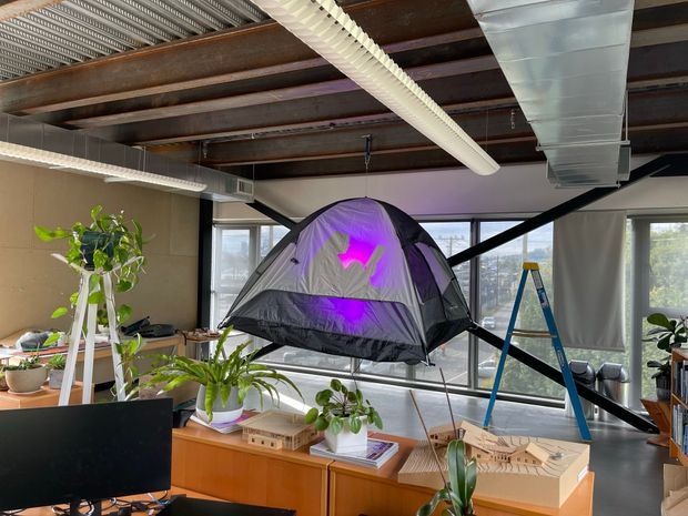 An industrial-style creative studio or office with exposed steel beams, corrugated metal ceiling, suspended linear lighting, and visible ductwork. Large windows provide abundant daylight. A suspended camping tent with purple interior lighting is the central feature, surrounded by desks, architectural models, books, computer equipment, and indoor plants.