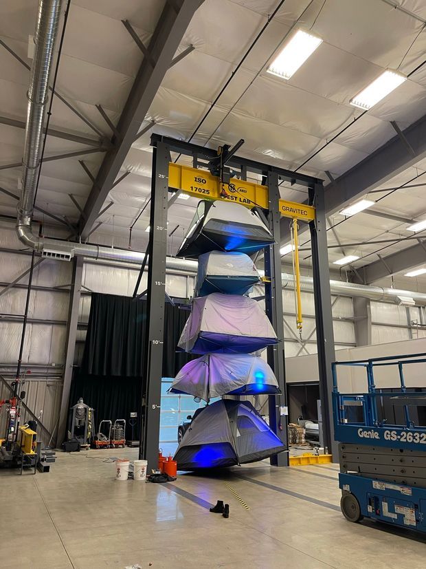 Industrial warehouse or testing facility interior with exposed steel framing, suspended insulated ceiling panels, ductwork, bright fluorescent lighting, and a tall yellow-and-grey test lab gantry. Several large fabric-covered test objects are stacked vertically within the rig, with an elevated work platform visible at right.