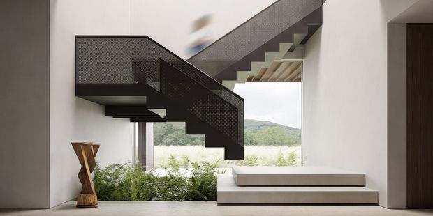 A contemporary interior stairway with a dramatic suspended zigzag staircase and black perforated metal balustrade. White walls, pale flooring, a low built-in bench or platform, indoor planting, and large glazing create a minimalist composition, with a green hillside visible outside. A sculptural timber hourglass-like object sits at the left.