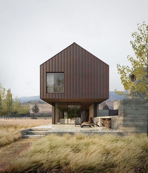 A contemporary residential house set in a dry, open landscape, photographed from the front. The compact upper volume has a steep gable roof and dark vertical metal cladding, with a large window on the facade. It is elevated over a glazed ground-floor living area and sheltered outdoor terrace. Pale concrete paving, a built-in outdoor fireplace, warm timber accents, and extensive ornamental grasses surround the home. A mountain or rural valley landscape is visible in the background.