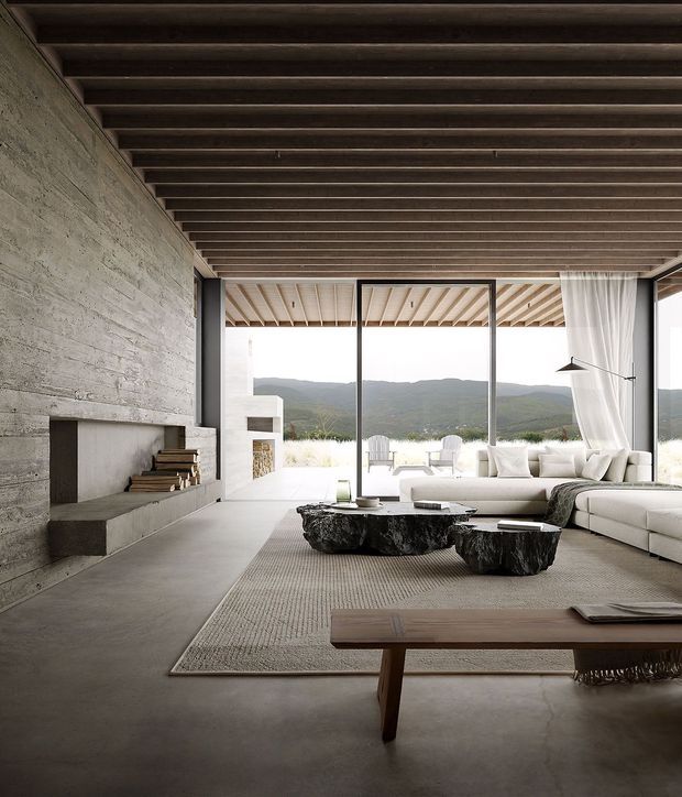 A spacious, refined living room with a strong indoor–outdoor connection. The room features a full-height rough concrete feature wall with a minimalist fireplace and integrated wood storage, a dark exposed timber-beam ceiling, polished concrete flooring, expansive black-framed glazing, and sheer curtains opening to a terrace and mountain landscape. A pale sectional sofa, textured neutral rug, sculptural dark stone coffee tables, and a simple timber bench create a warm yet pared-back interior.
