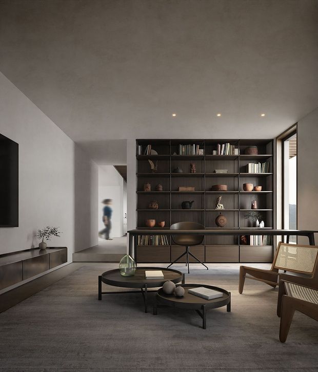 A refined contemporary living room with a large dark built-in bookshelf and display wall, low black nesting coffee tables, a streamlined media console, and timber lounge chairs with woven cane backs. The room has a restrained grey, charcoal, and timber palette, smooth plaster walls, recessed ceiling lights, and a tall glazed opening beside the shelving. A hallway extends behind the room.