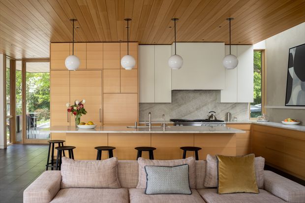 Open-plan kitchen viewed from the living area, featuring extensive light timber cabinetry, a large island with seating, white upper cabinets, a stone-look splashback, integrated appliances, globe pendant lights, and a timber-lined ceiling. A large sectional sofa occupies the foreground, with glazed doors and windows bringing in views of greenery.