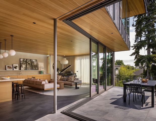 A refined contemporary indoor-outdoor living space with a warm timber-lined ceiling extending from the interior to the covered terrace. Full-height glazed sliding doors open the living room to a paved outdoor dining area. Inside are a neutral sectional sofa, a grand piano, sculptural pendant lights, and a streamlined kitchen with integrated cabinetry. The terrace has a dark dining table and chairs overlooking a leafy suburban setting.