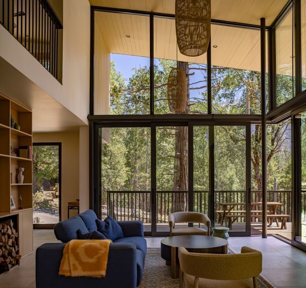 A warm, contemporary double-height living room with expansive floor-to-ceiling glazed doors and windows opening to a timber deck surrounded by trees. The room includes a deep blue sofa, rounded lounge chairs, a low circular coffee table, built-in shelving, sculptural woven pendant lights, and a black metal balcony railing on the upper level. Large dark-framed glazing provides a strong connection to the natural setting.