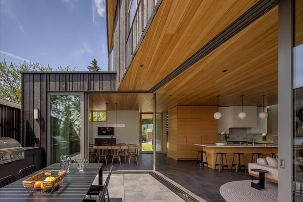 A contemporary indoor-outdoor entertaining area viewed from a covered patio or terrace. The image shows a timber-lined soffit and ceiling, dark vertical exterior cladding, expansive glazed sliding doors, a dining area, and an open-plan kitchen with a long island and stools. A built-in outdoor grill and dining table occupy the foreground, while the interior continues seamlessly beyond the open glazing.