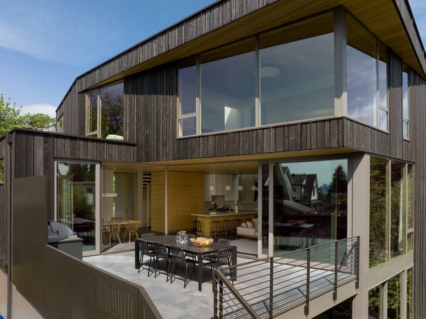Exterior view of a contemporary multi-level house with dark vertical timber cladding, extensive floor-to-ceiling glazing, projecting upper-level volumes, and a covered outdoor terrace. The terrace includes dining furniture and connects visually to an open-plan kitchen and living area behind the glazing. A metal balustrade and broad concrete terrace surface define the outdoor space.