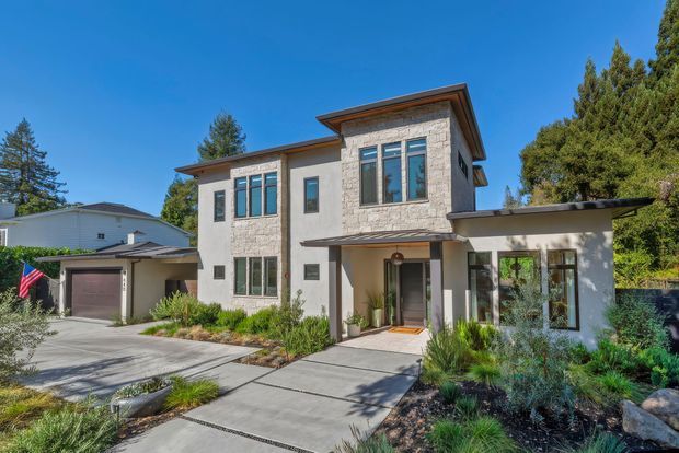 Exterior view of a contemporary multi-storey residence with a pale rendered facade, prominent light stone-clad upper wall, dark metal low-pitched roof edges, large framed windows, and an attached garage. Concrete paths and restrained drought-tolerant landscaping lead to the recessed entry.