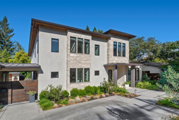 A contemporary two-storey residence viewed from the street, set among mature trees and landscaped planting. The facade combines smooth light rendered walls with pale natural stone cladding, dark-framed rectangular windows, and broad low-pitched roof planes with timber-toned soffits. A covered carport and timber garage door sit to the left, while a paved driveway and sheltered entry define the foreground.