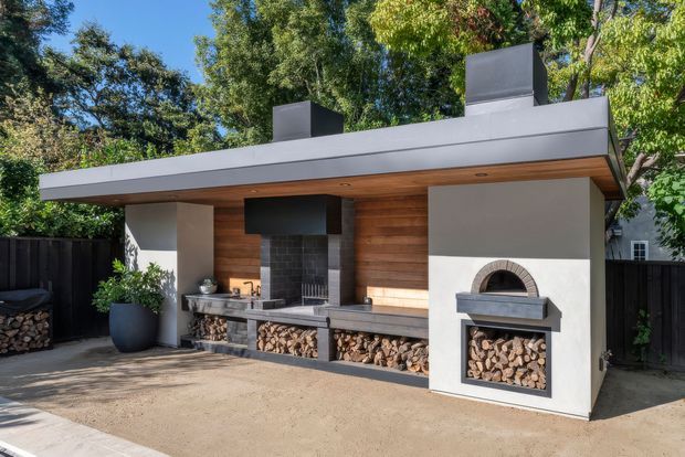 A contemporary outdoor entertaining area with a flat-roofed structure, integrated wood-fired pizza oven, large open fireplace, dark brick fire surrounds, timber-lined ceiling and wall, white rendered walls, and neatly stacked firewood storage. The setting is landscaped and tree-lined, with a paved or concrete forecourt.