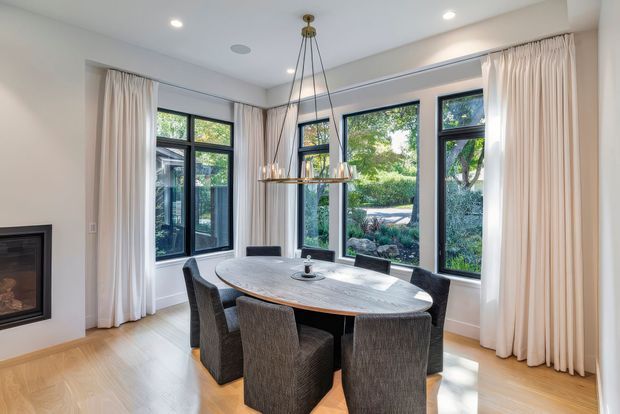 Bright, refined dining room with a large oval stone-topped dining table surrounded by upholstered chairs. Floor-to-ceiling windows with dark frames and full cream curtains overlook a landscaped garden, while a linear pendant light hangs above the table. Light timber flooring, white walls, recessed lighting, and a built-in fireplace create a warm contemporary setting.