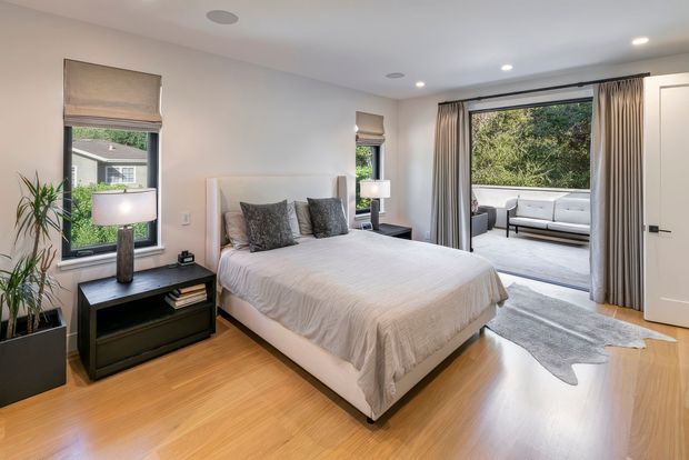 A bright, contemporary bedroom with a large upholstered bed, neutral bedding, dark bedside furniture, and table lamps. The room has warm timber flooring, pale walls, roller blinds, and full-height curtains framing an open doorway to a furnished outdoor or adjoining lounge area. Recessed ceiling lights and indoor plants complete the clean, comfortable design.