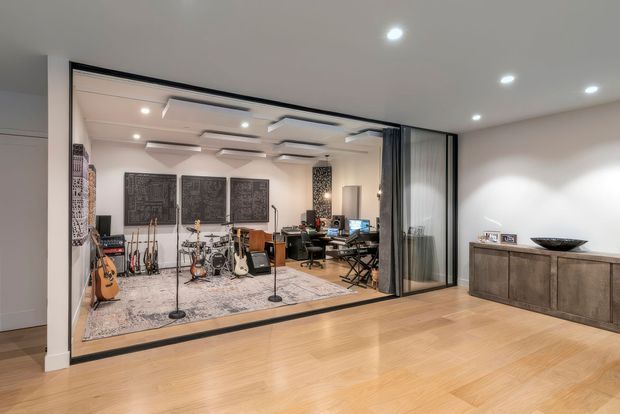 A contemporary open-plan interior with a polished timber floor and a large glazed, black-framed partition enclosing a music studio or rehearsal room. The studio contains guitars, microphones, keyboards, computer equipment, acoustic wall panels and patterned rugs. In the foreground, a minimalist console cabinet with decorative objects sits against a white wall. Recessed ceiling lights and layered acoustic ceiling panels create a clean, refined look.