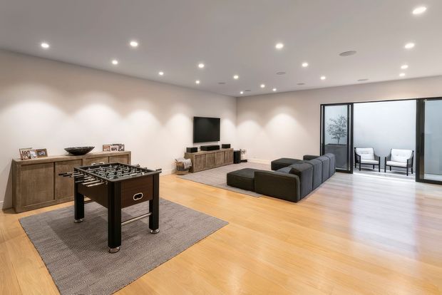 Spacious contemporary media and recreation room with pale timber flooring, a dark foosball table on a large rug, sectional seating facing a wall-mounted television, low timber cabinetry, recessed ceiling lighting, and full-height glazing opening to a small outdoor seating area.