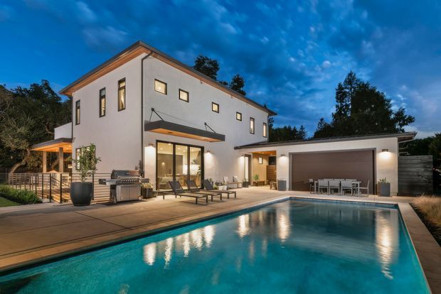 Twilight exterior view of a contemporary two-storey white-rendered residence with a low-pitched roof, large glazed openings, covered outdoor entertaining areas, an adjoining open garage or pavilion, and a rectangular swimming pool with surrounding paving and landscaping.